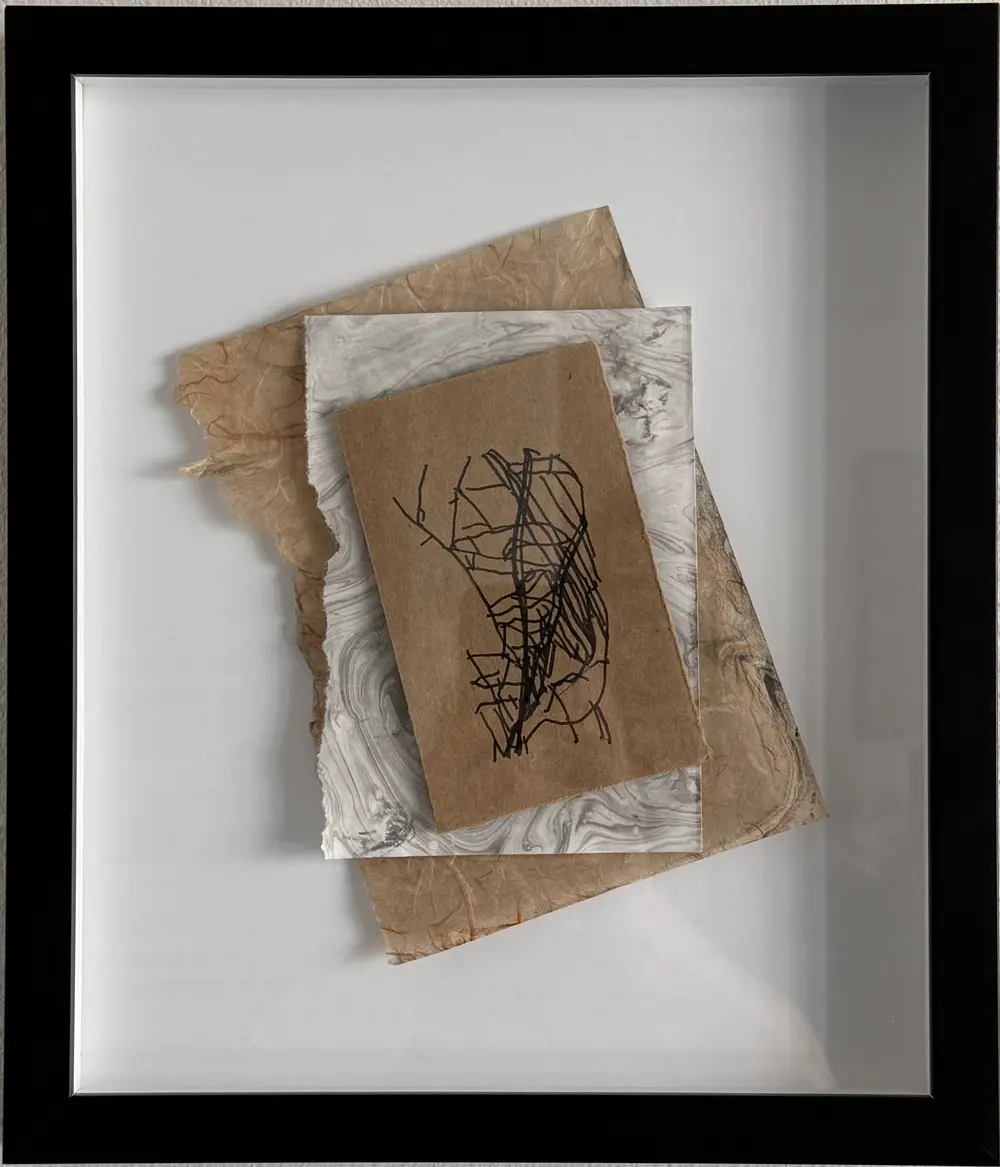 Fracture in a black shadow-box frame: a kraft card drawn with tangled, wobbly black lines, layered over grey-and-white marbled paper and torn tan fibrous paper