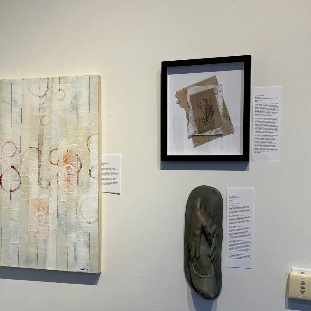 Gallery wall in the Memory Lane exhibition: Fracture in its black frame beside its printed story label, next to a large textured white panel with red circles, above a bronze relief
