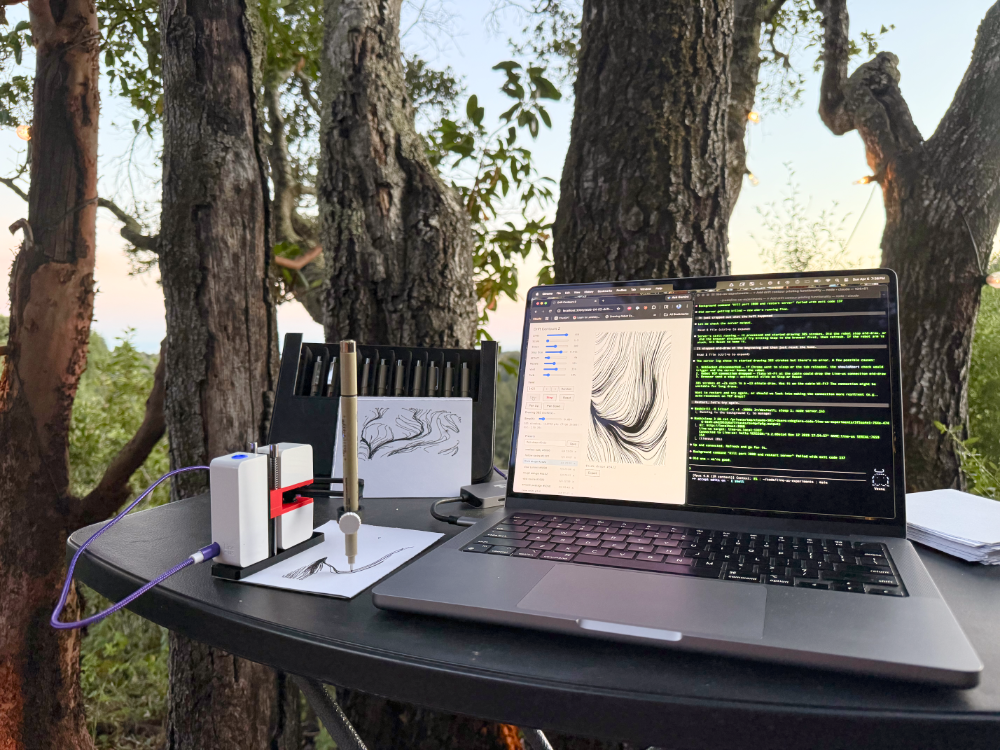 Outdoor workstation at sunset overlooking a redwood ravine — MacBook running Claude Code and the Drift Contours p5.js interface, Line-us mid-drawing, sketches and pens on the table